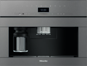 Integrated Miele coffee machine with chrome brew head, digital clock display, and glass carafe visible on the left within a dark-gray housing.