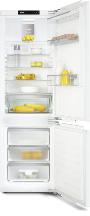 Open white refrigerator with shelves containing lemons, eggs, milk, juice, peppers, and two yellow zucchinis in the bottom drawer
