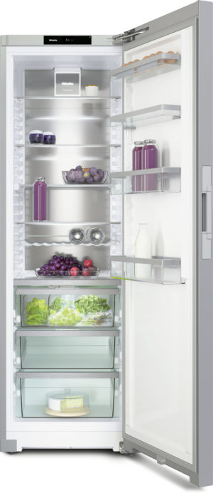 Open stainless steel refrigerator with glass door, showing organized shelves: bottles of juice, jars, a bowl of fruit, dairy on top shelf, crates of grapes and greens in drawers, and a cake on the bottom shelf; interior lighting highlights the organized storage.