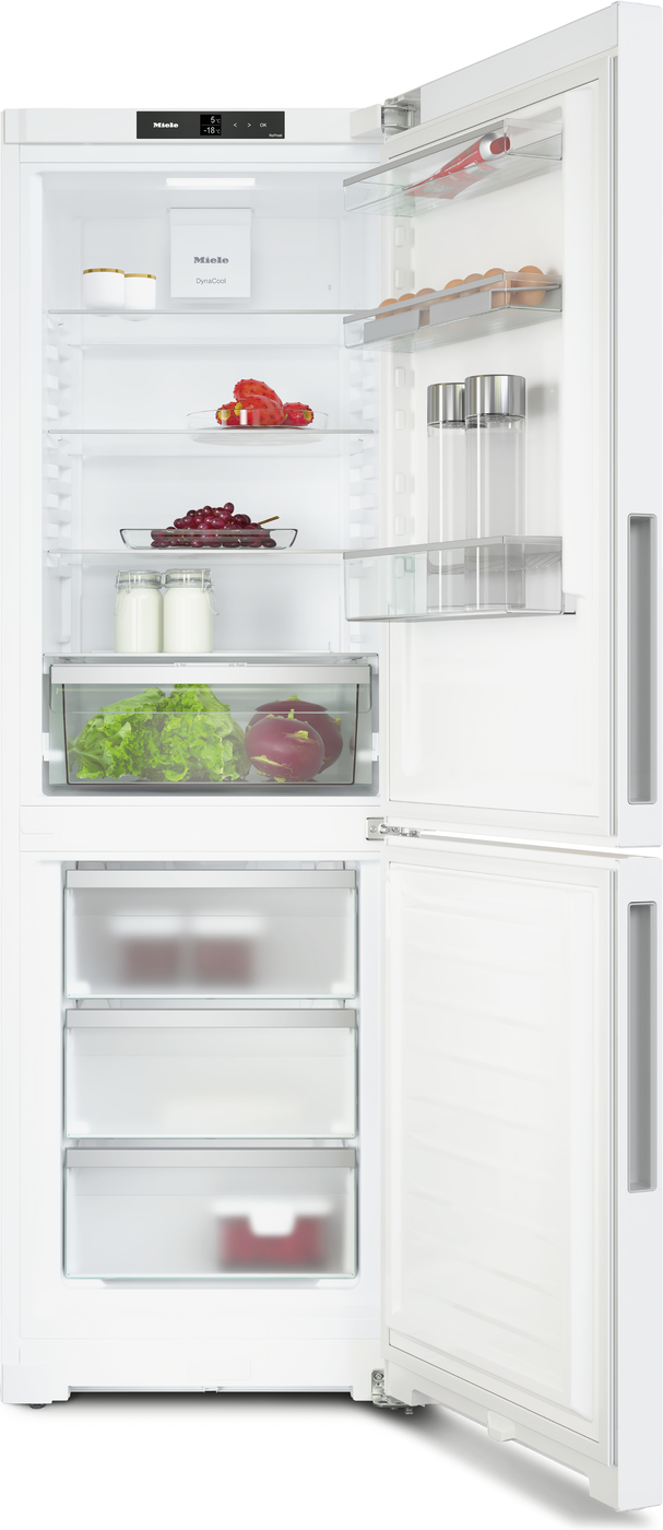 Open white refrigerator with upper fridge and lower freezer. Shelves hold milk, grapes, lettuce, tomatoes, eggs, and jars; interior light on.