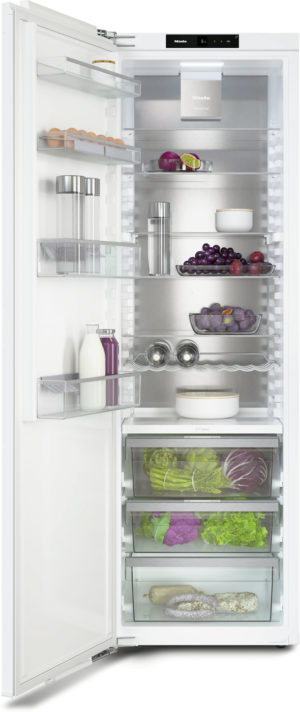 Open white refrigerator showing shelves with eggs, dairy, fruit, and vegetables; bottom drawers contain greens and root vegetables; interior lights on, modern Miele appliance visible at top