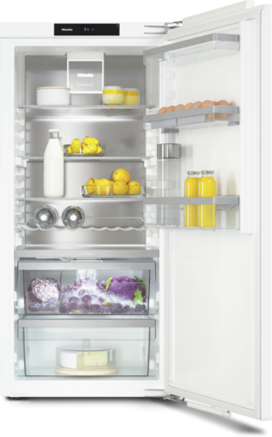 Open white refrigerator with stainless interior showing organized shelves: milk bottle, eggs, citrus fruits, juice bottles, cheese wheel, vegetables, and a cake on the bottom shelf.