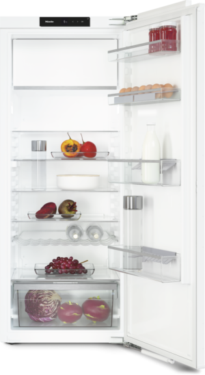 Open refrigerator door revealing a clean white interior with a glass top shelf, milk bottle, eggs, jars, fresh fruit (grapes, tomatoes, peppers), and vegetables (cabbage) on clear shelves and drawers.