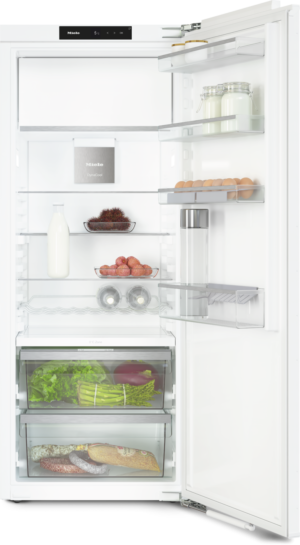 Open white refrigerator showing shelves with milk bottle, eggs, vegetables, jars, and sausage; bottom freezer.