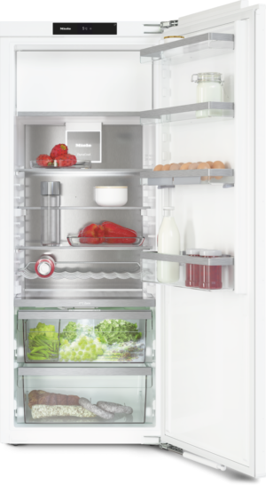 Open refrigerator door showing organized shelves: dairy and jars on the upper shelves, peppers and fruit mid-level, greens and grapes in the drawer, and bread and sausages in the bottom bin.