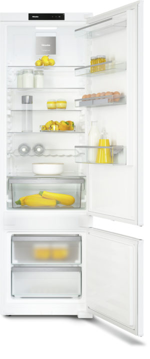 White refrigerator with doors open, showing organized shelves: lemons, milk, eggs, jars, yellow condiments, and a bottom freezer with yellow squash.
