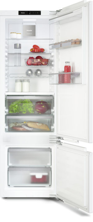 Open white Miele refrigerator with bottom freezer; shelves hold milk, tomatoes, lettuce, bread, sausages, eggs, jars, and a bottle. Fruit basket visible on a shelf.