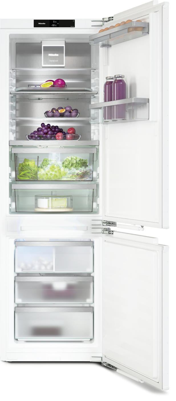 White French-door style refrigerator with top freezer, open doors, showing organized shelves of fruits, vegetables, and bottles on the door shelves; bottom freezer drawers visible.