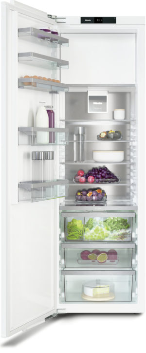 Open refrigerator with glass shelves, displaying eggs, milk, juice, produce, fruits, cheese, and a cake; interior lights on.