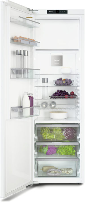 Open white refrigerator with glass shelves holding eggs, dairy bottles, bowls, fruit, and vegetables; bottom freezer drawers reveal greens and roots.