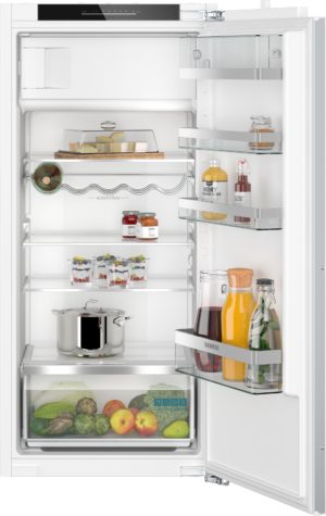 Open white refrigerator with organized shelves: cheese on a cutting board, jars, condiments, bottles, and a bottom fruit/vegetable drawer visible.