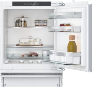 Open white refrigerator showing dessert cake and fruit cups on the left shelf, and jars of preserves on the right shelf.
