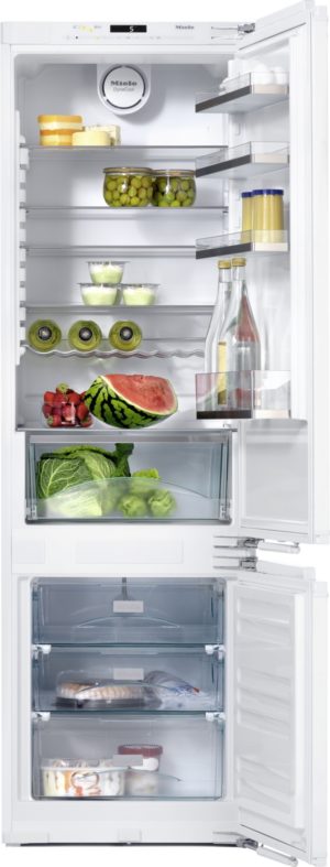 Open white refrigerator filled with food: cheeses, jars of olives, bottles, fresh produce, and a watermelon half on the shelves and in the crisper drawers