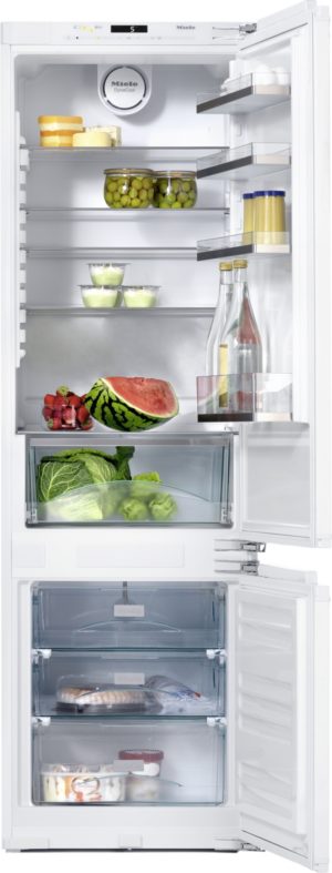 Open white refrigerator stocked with foods: cheeses on top shelf, jars of olives, bottles and condiments on the door shelves, a watermelon half and berries on the middle shelf, greens and lettuce in the crisper, and a freezer drawer with seafood and desserts visible through the transparent drawers.