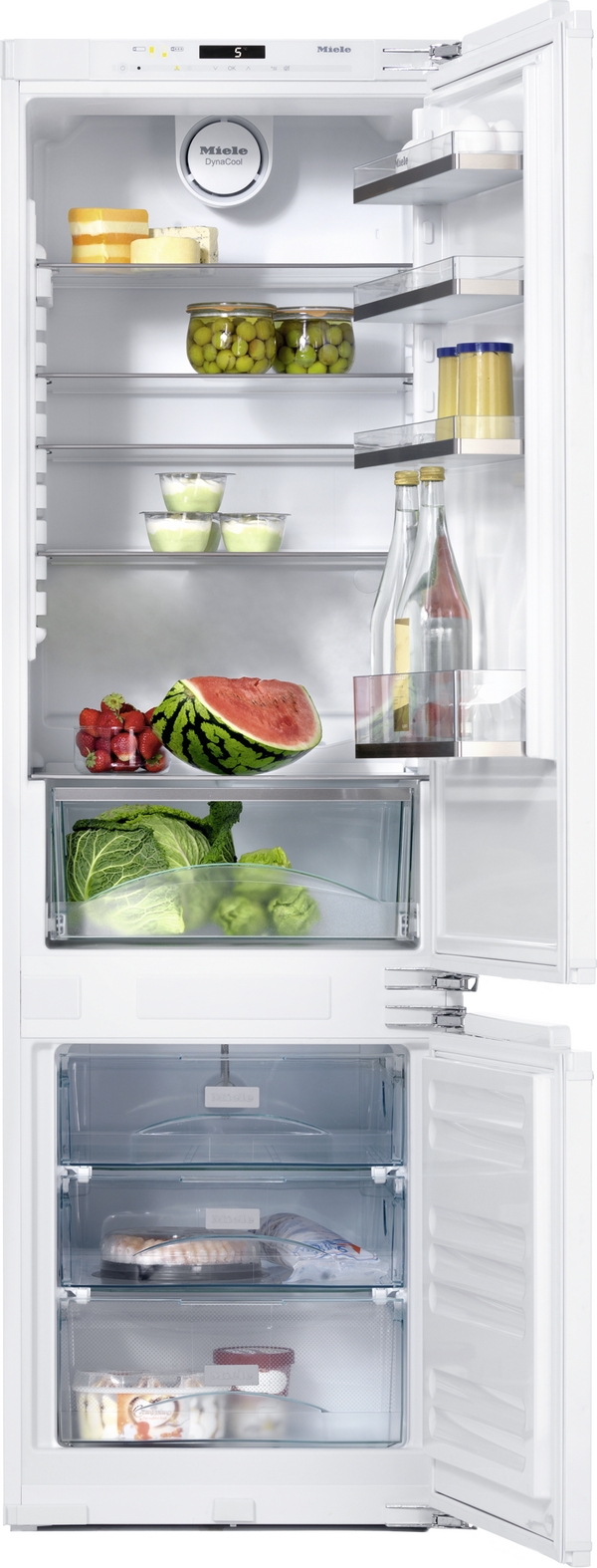 Open white refrigerator stocked with foods: cheeses on top shelf, jars of olives, bottles and condiments on the door shelves, a watermelon half and berries on the middle shelf, greens and lettuce in the crisper, and a freezer drawer with seafood and desserts visible through the transparent drawers.
