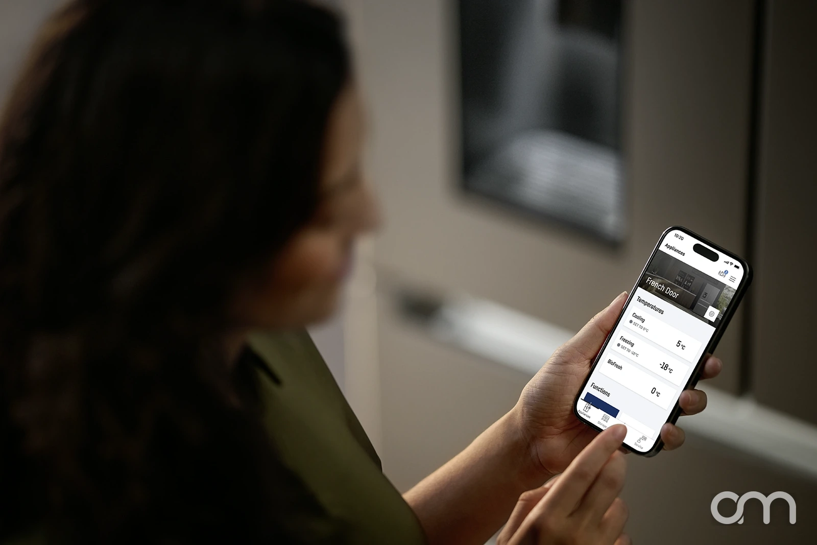 Femme consultant l'application Liebherr SmartDevice sur son smartphone devant son réfrigérateur, températures et réglages affichés à l'écran : conseil en électroménager connecté par Arts & Ménagers, Neuchâtel